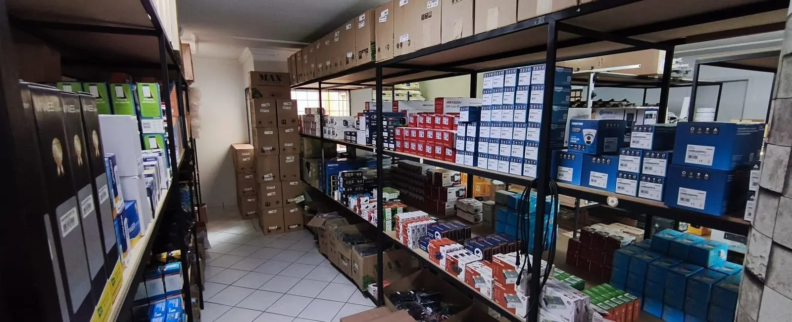 Corredor do depósito da Capital Distribuidora com prateleiras cheias de caixas de câmeras, gravadores, fontes e acessórios de segurança eletrônica
